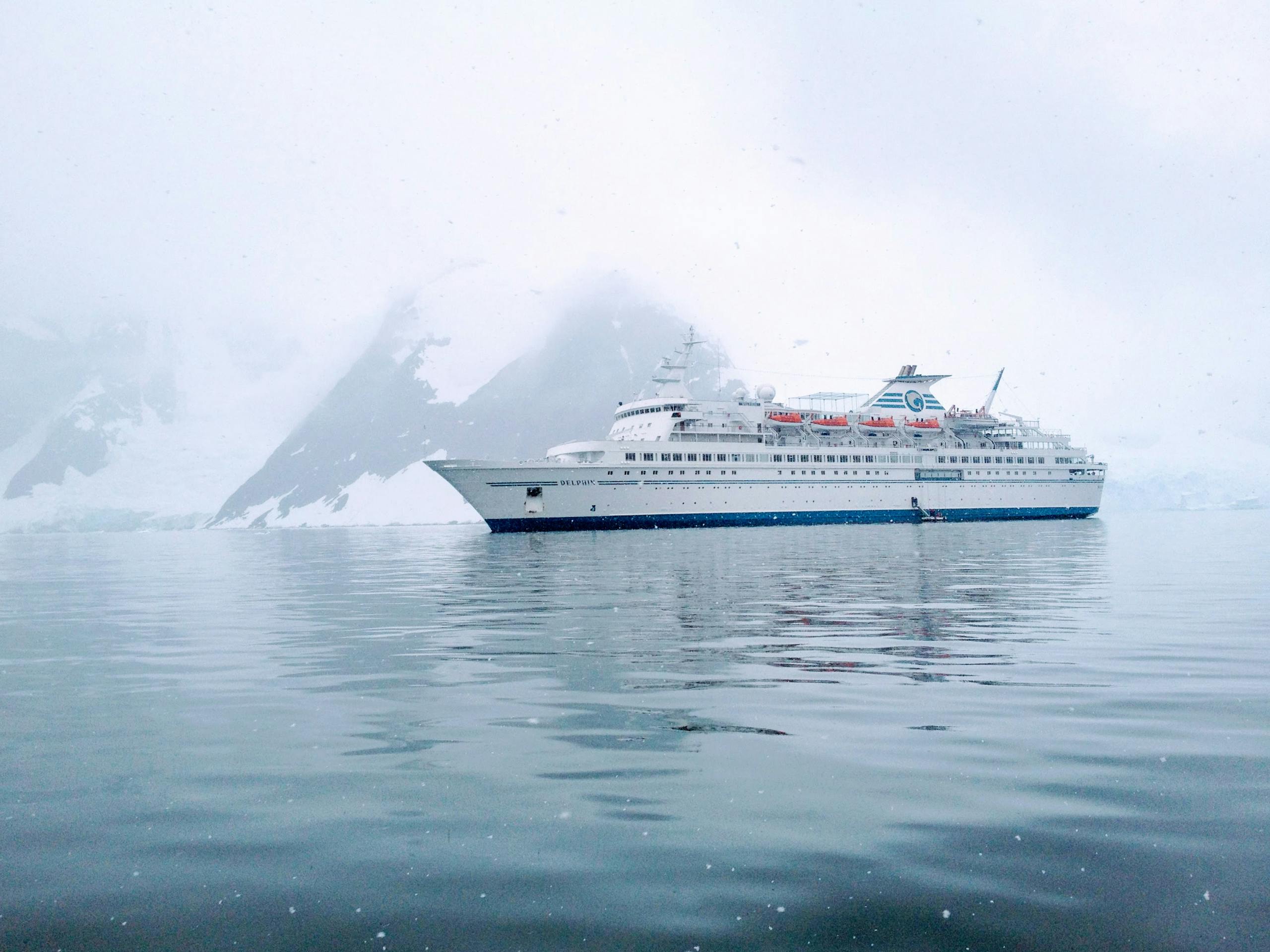 A cruise ship sails calmly through the icy waters of Antarctica, surrounded by snow-capped mountains.cruise travel advisor Simply Adventurous Travel